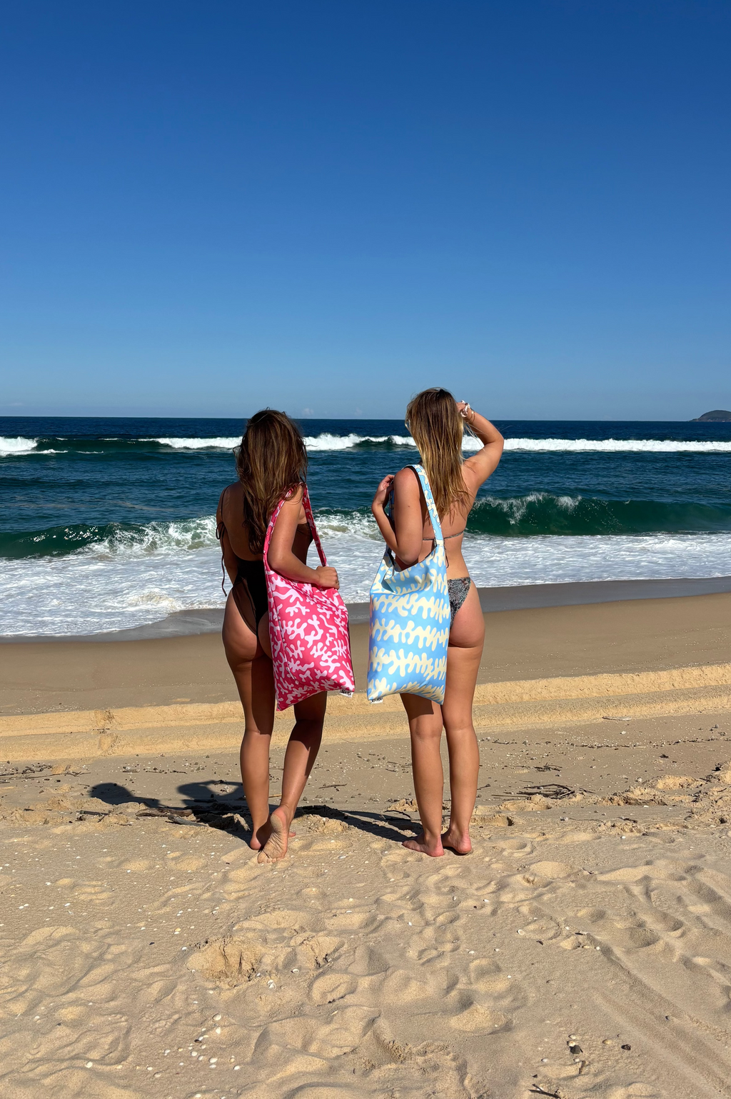 Two women on the beach carrying SEA PEA beach bean bags in Blue Ripple and Pink Coral, looking out at the ocean.