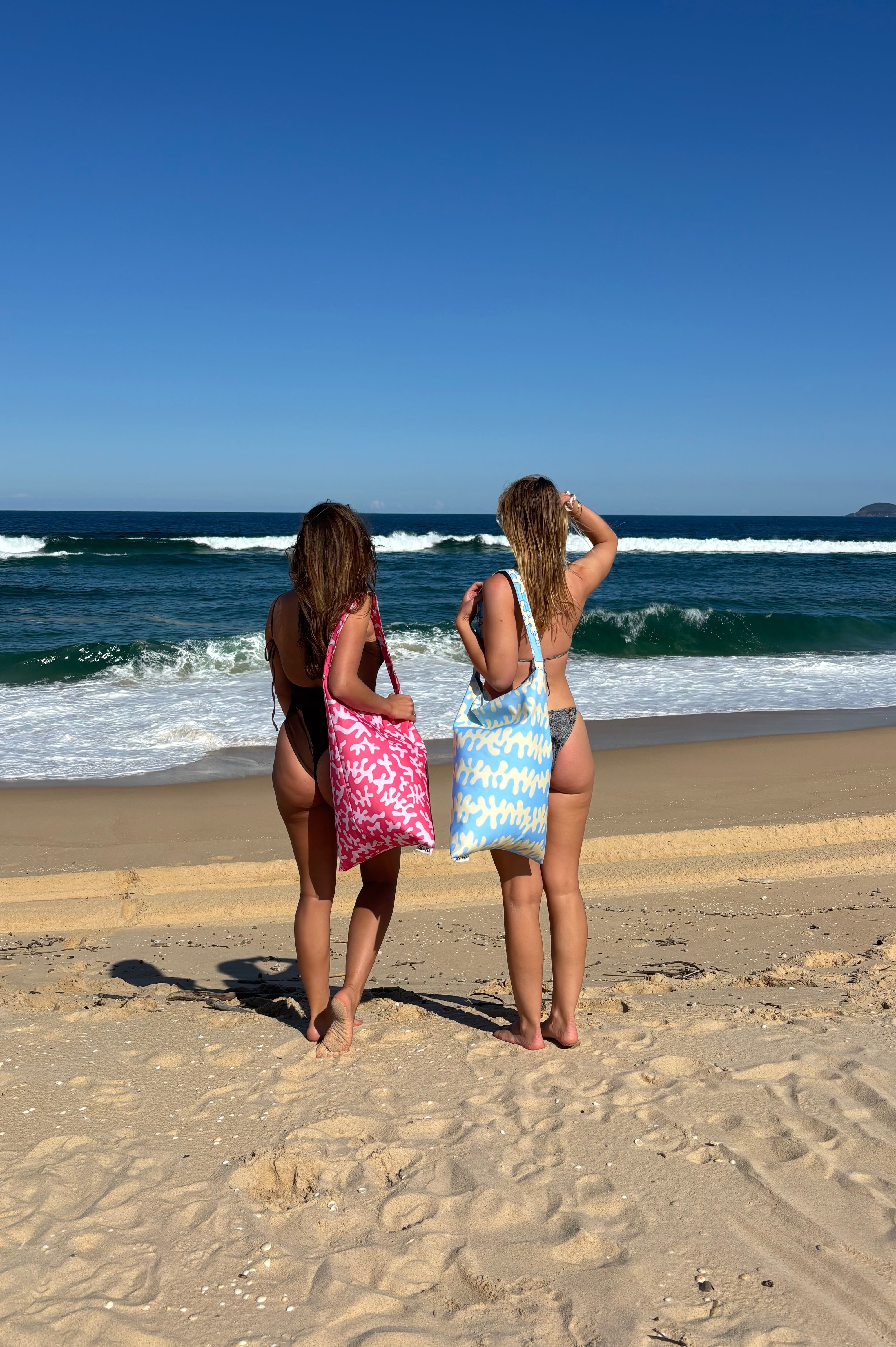 Two women on the beach carrying SEA PEA beach bean bags in Blue Ripple and Pink Coral, looking out at the ocean.