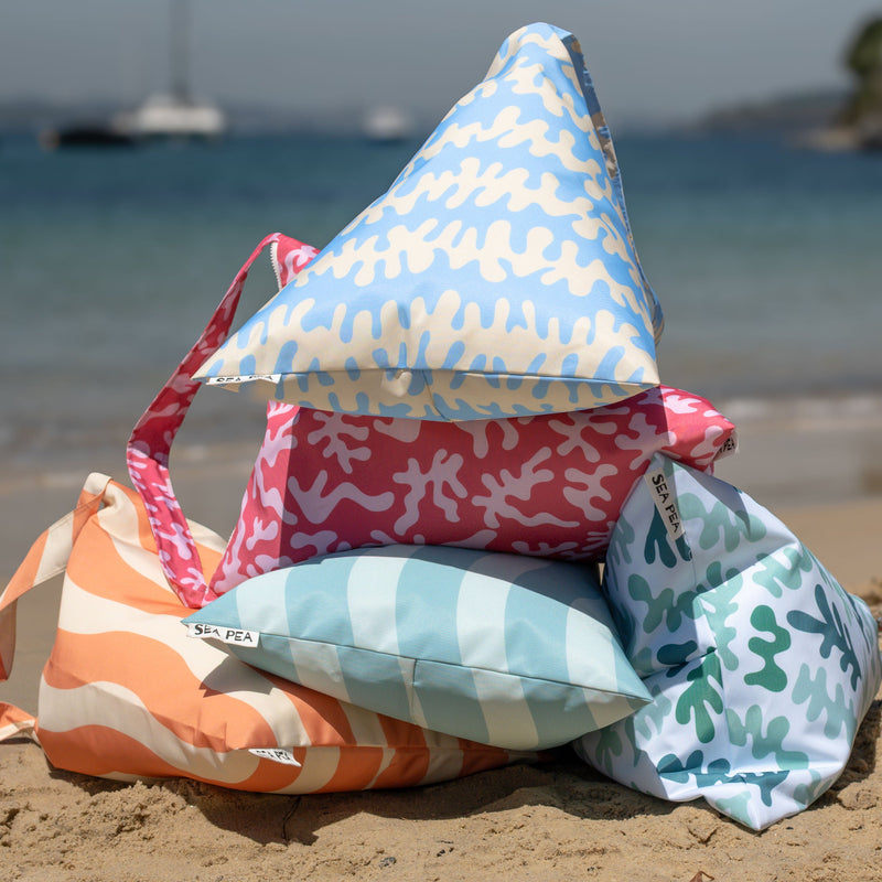 Colorful beach bag SEA PEAs stacked on a sandy beach with ocean and sailboat in the background